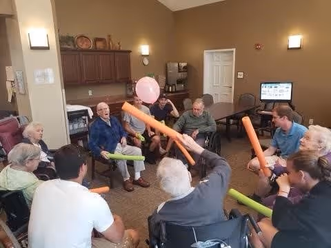 A group of elderly people and caregivers sitting in a circle in a room, engaging in a balloon volleyball activity using colorful foam noodles. The room has beige walls, a table with a computer, and cabinets in the background.