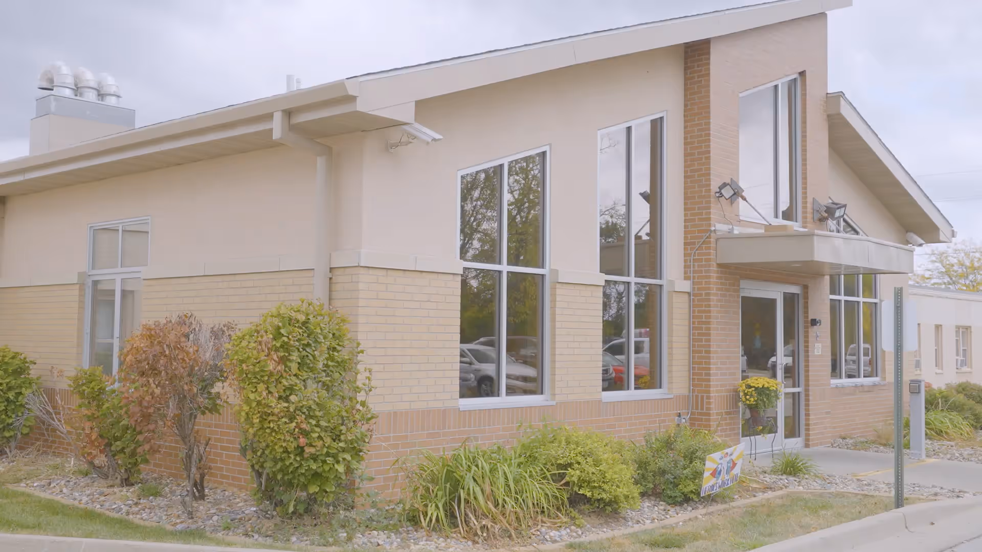 Front entrance of a low-rise brick and stucco senior living building with large windows, shrubs, and a sidewalk.