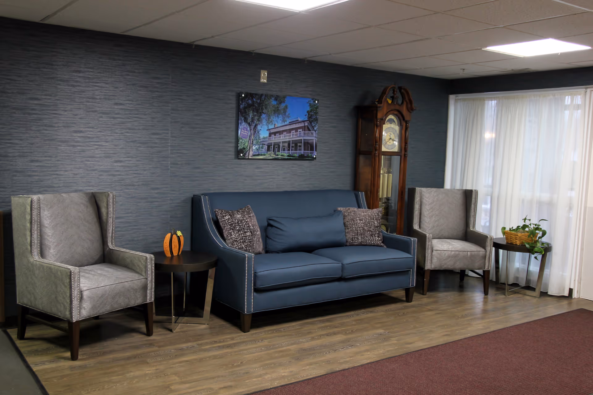 A seating area with a blue sofa, two gray armchairs, side tables, a grandfather clock and framed art in a common room.