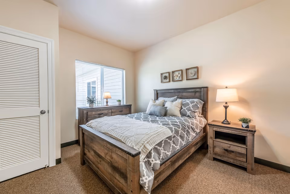 A cozy bedroom with a wooden bed, matching nightstand and dresser, a lamp, and a window.