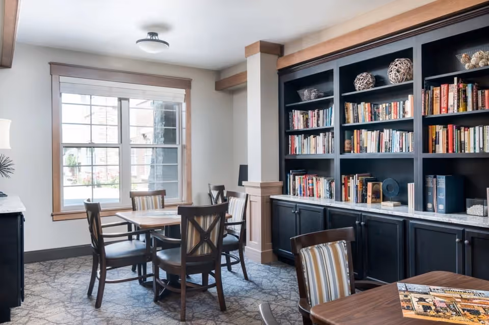 A cozy reading or common area with a large window letting in natural light, several wooden chairs with striped cushions around wooden tables, and a built-in black bookshelf filled with books and decorative items.