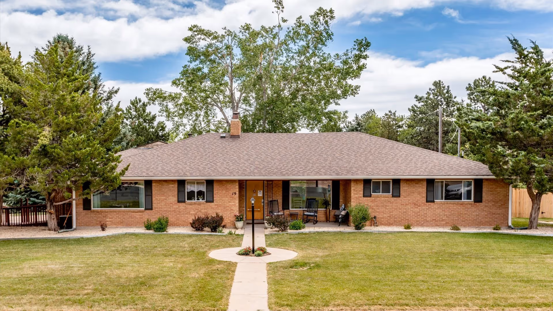 Single-story brick building with a brown shingled roof, surrounded by green grass and trees under a partly cloudy sky. A concrete path leads to the front door, which is flanked by windows and small shrubs. There are two rocking chairs on the front porch.