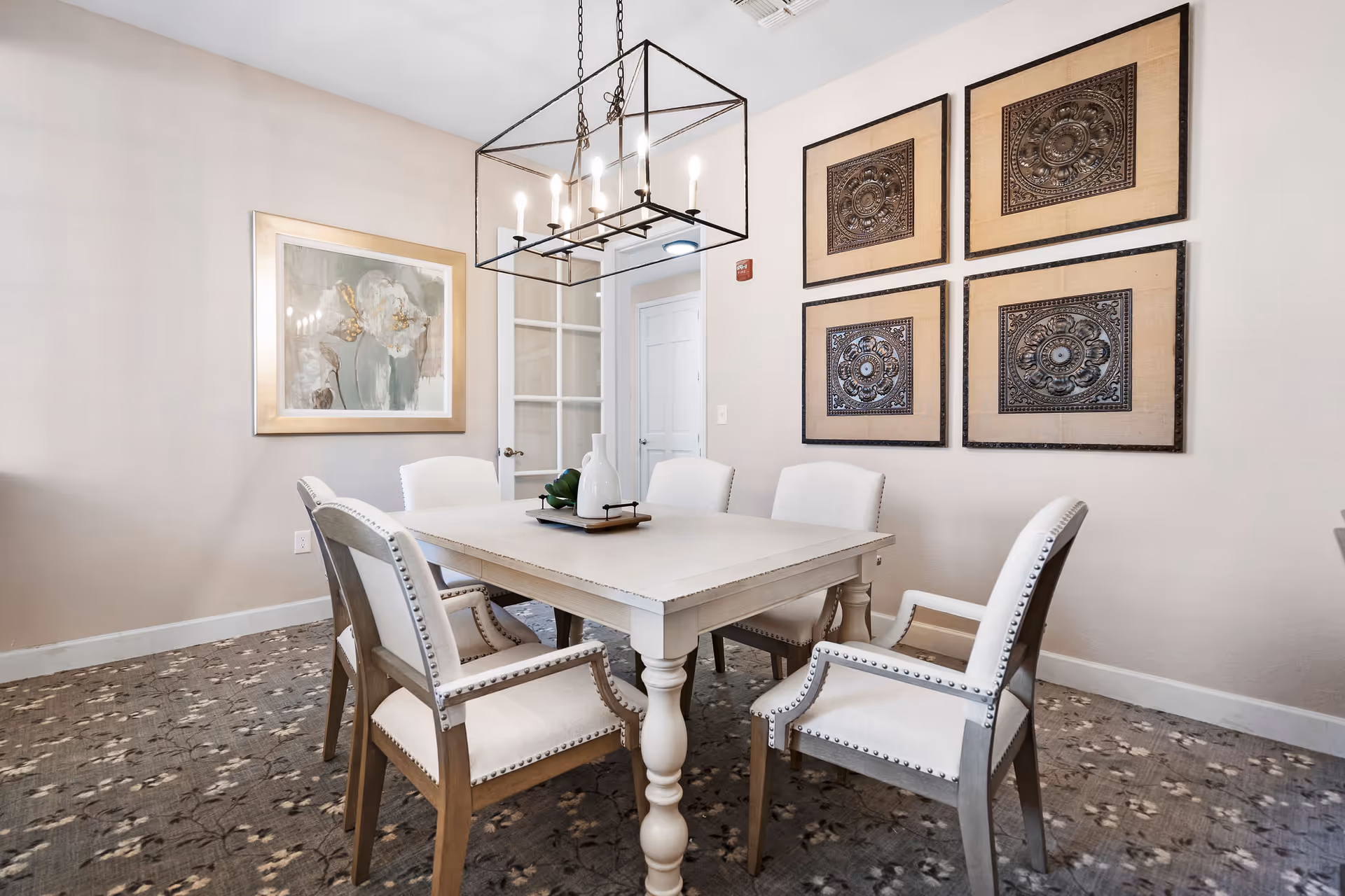 A dining room with a white wooden table surrounded by six upholstered chairs with nailhead trim. Above the table hangs a rectangular chandelier with exposed bulbs. The walls are decorated with a framed floral painting and four square decorative metal wall art pieces. The floor is carpeted with a patterned design, and a glass-paneled door is visible in the background.