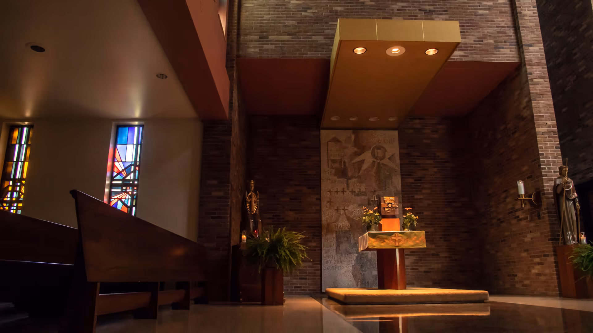 Interior view of a chapel with wooden pews on the left, stained glass windows, a central altar with a cloth featuring a cross, plants on either side, statues, and brick walls.