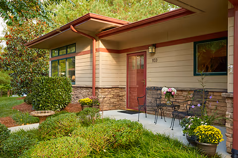 Exterior view of a single-story residential unit with beige siding and stone accents, a red door labeled 103, a small patio with a black metal table and two chairs, and potted flowers. The surrounding area features green shrubs, a birdbath, and trees.