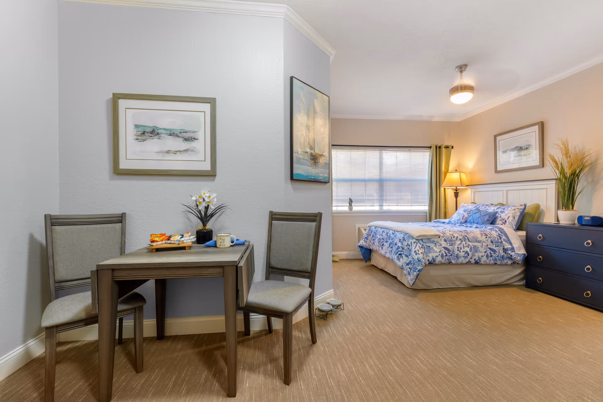 A cozy senior living bedroom with a bed dressed in blue and white floral bedding, a dark blue dresser with a potted plant and clock on top, a floor lamp, and a window with blinds and green curtains. Adjacent to the bedroom area is a small dining table with two chairs, a vase with flowers, snacks, and a coffee mug. The walls are decorated with framed artwork.