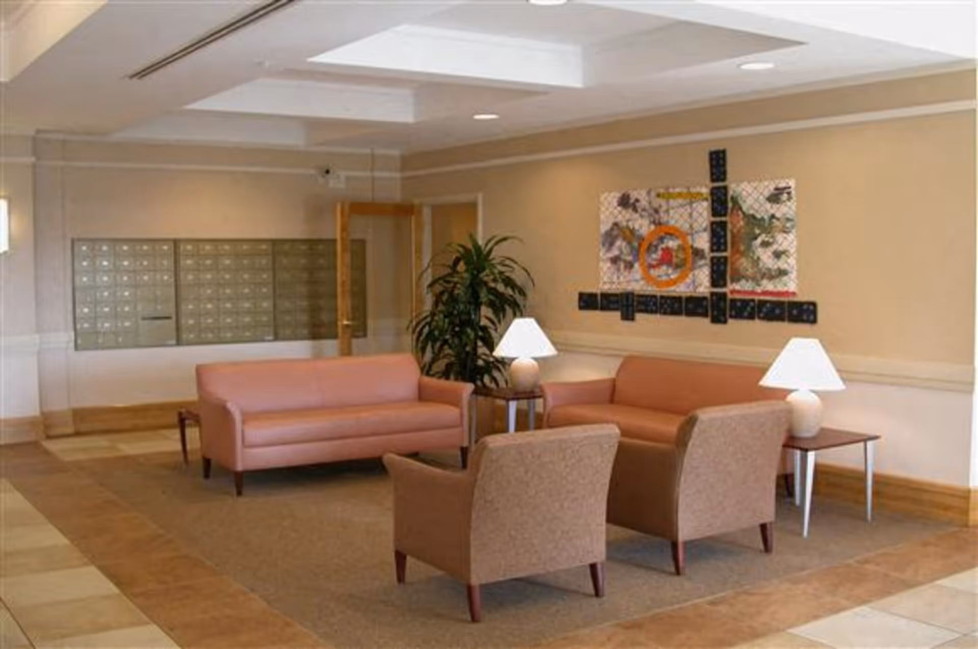 A seating area in a senior living facility lobby with two pink sofas, two brown armchairs, two side tables each with a white lamp, a large potted plant, and a colorful abstract wall art. Behind the seating area is a wall of mailboxes and a wooden door.