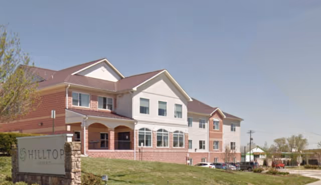 Exterior view of a multi-story senior living facility named Hilltop, featuring a combination of brick and white siding with multiple windows and a sloped roof, situated on a grassy area with a sign in front.