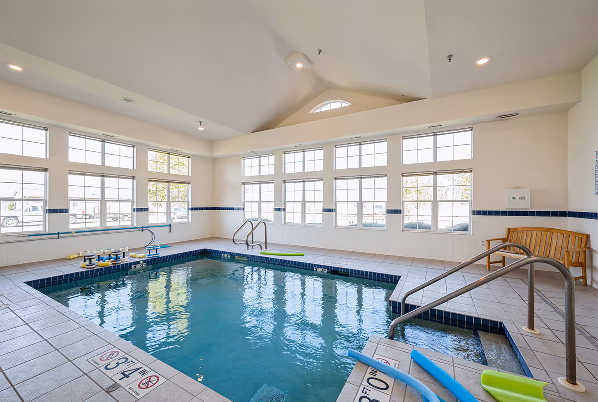 Indoor community swimming pool with handrails, pool noodles, and large windows letting in natural light.