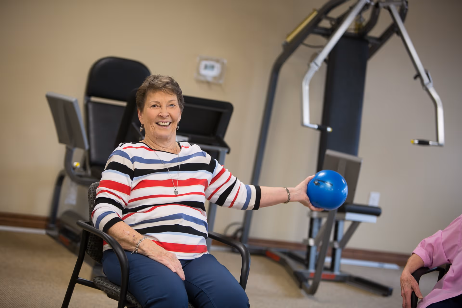 An elderly woman sitting on a chair in a fitness room, smiling and holding a small blue exercise ball with one hand. Exercise equipment is visible in the background.