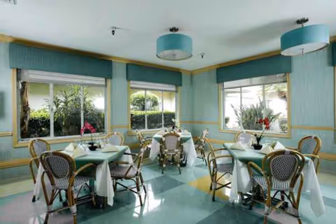 Pastel teal dining room with round tables, wicker chairs, and large windows.