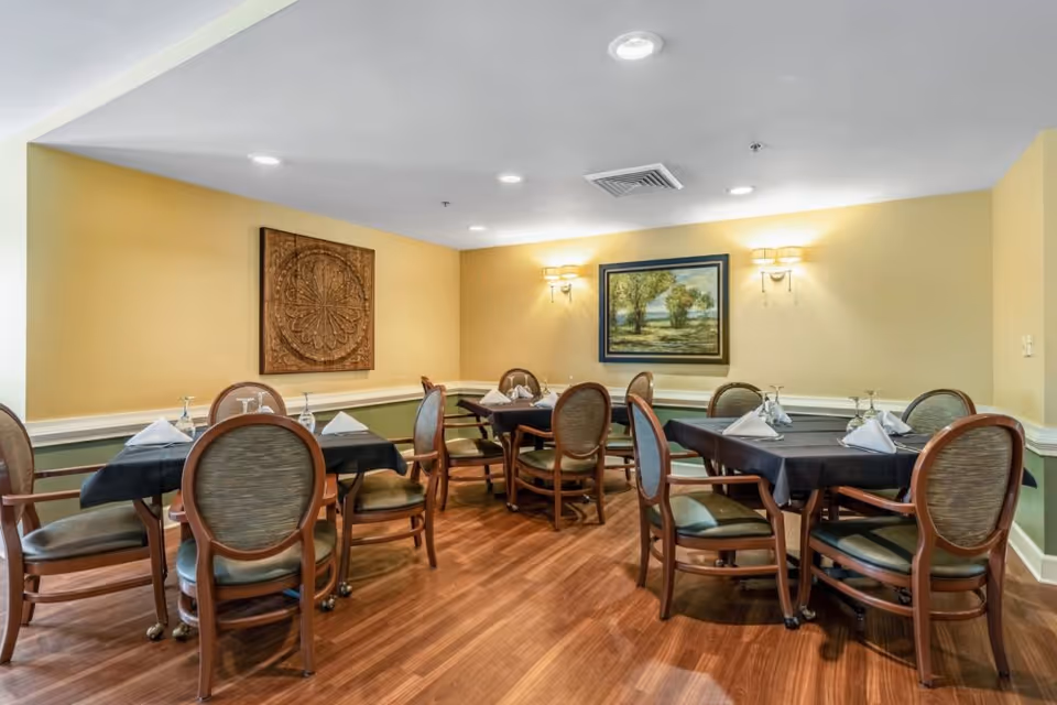 Dining room with several tables covered with black tablecloths, each set with folded white napkins and upside-down wine glasses. The room has wooden chairs with cushioned seats and backs, yellow walls with green wainscoting, wooden flooring, and wall decorations including a framed painting and a carved wooden panel.