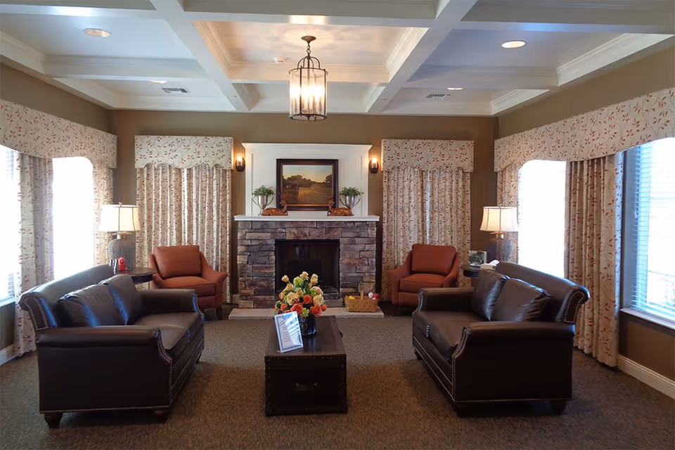 A cozy living room with two dark brown leather sofas facing each other across a wooden coffee table adorned with a flower arrangement. The room features a stone fireplace with a framed landscape painting above it, flanked by two small plants and two brown armchairs. The walls are painted brown and decorated with floral curtains covering the windows. Two table lamps with beige shades provide soft lighting, and a ceiling light fixture hangs above.
