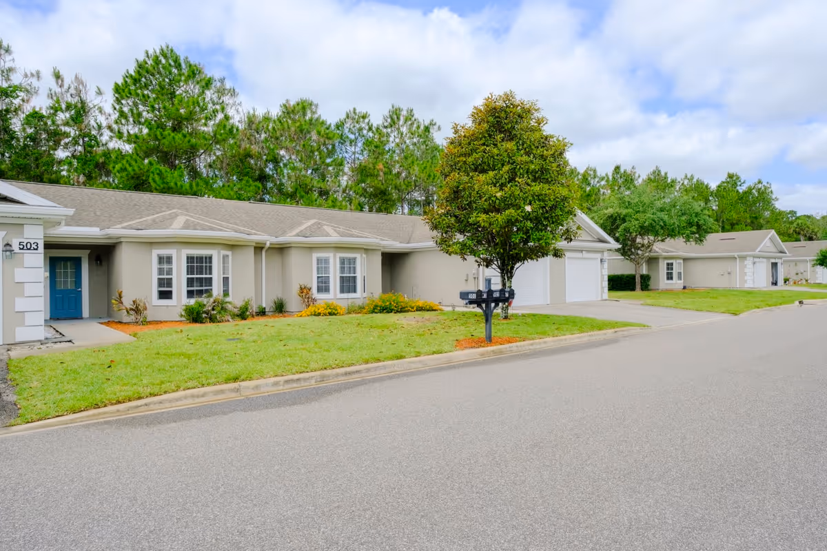 Single-story beige townhome-style units with garages, lawns, a mailbox and trees along a quiet street under a partly cloudy sky.