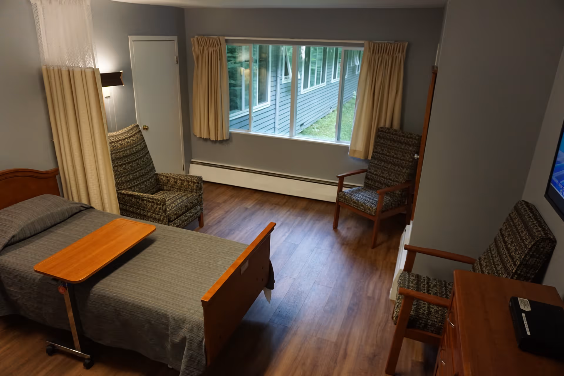 A nursing-home bedroom with a single bed and overbed table, three upholstered chairs, a dresser, and a large window.