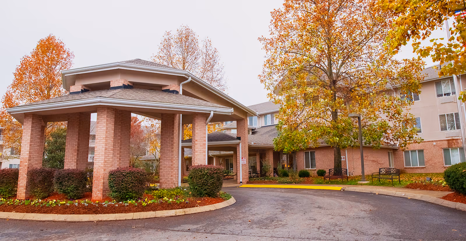 Front entrance of a brick senior living building with a covered porte-cochère and autumn trees.