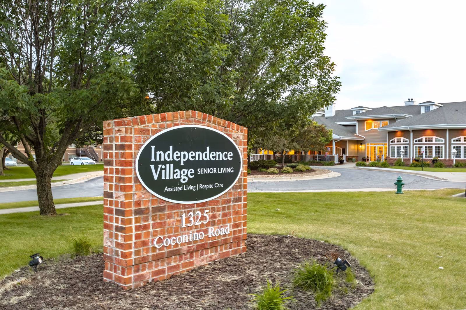 Brick sign for Independence Village Senior Living at 1325 Coconino Road with a senior living facility building and landscaped grounds in the background.