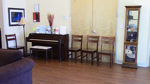 Interior room with wooden floor featuring a piano with a bench, four wooden chairs lined up against a wall with double doors, a tall glass display cabinet with decorative items, a floor lamp, and a framed picture on the wall.