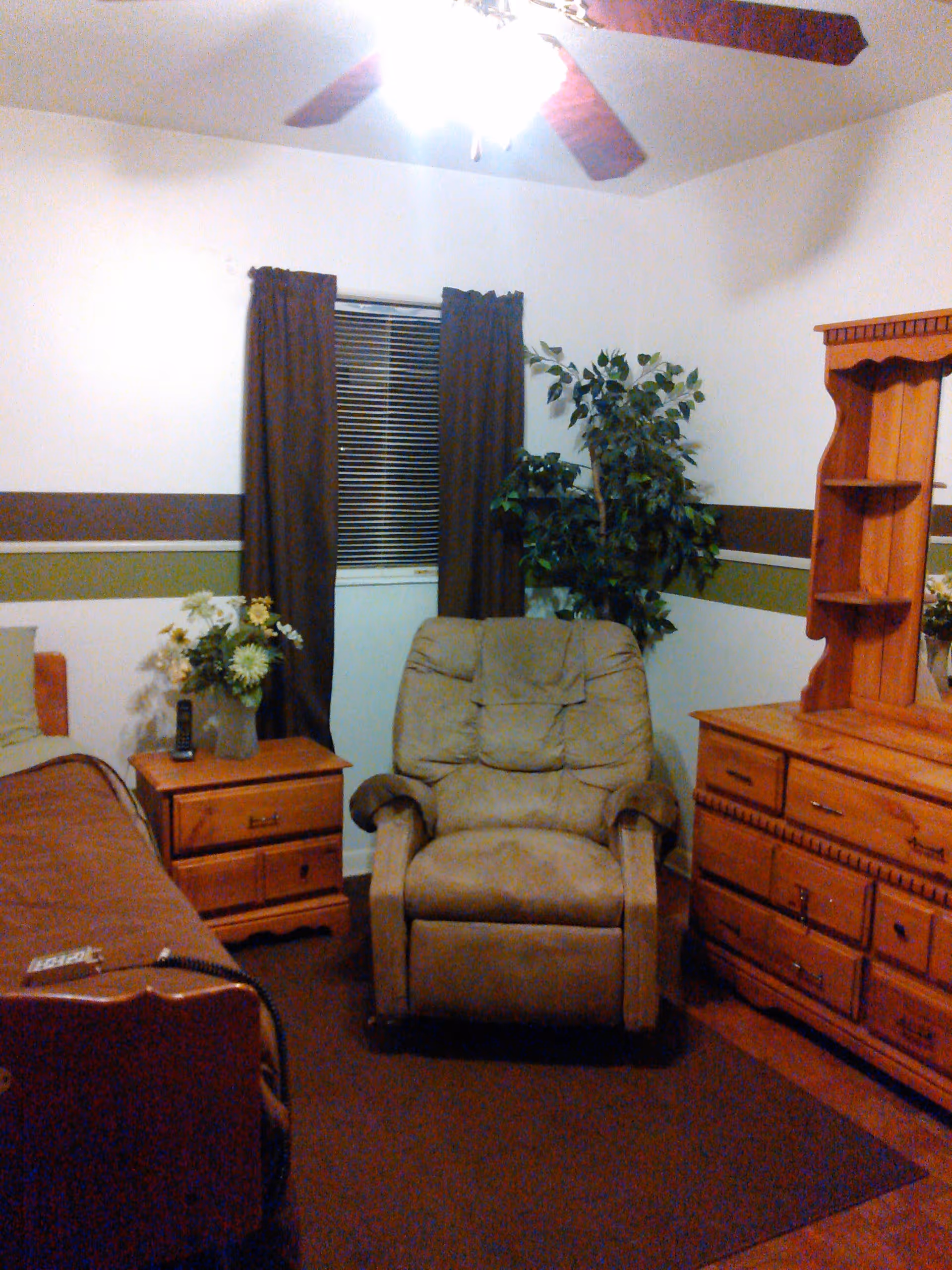 Bedroom with a beige recliner centered between a bed and wooden dresser, a window with dark curtains and a ceiling fan overhead.