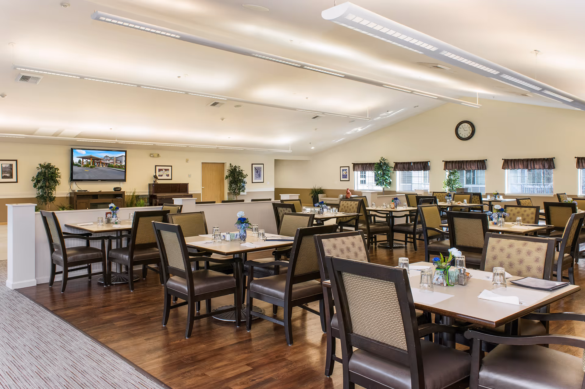 A spacious dining room with multiple tables and chairs arranged neatly. Each table is set with glasses, napkins, and small flower vases. The room has wooden flooring, large windows with valances, and plants placed around. A wall clock and a TV mounted on the wall are also visible.