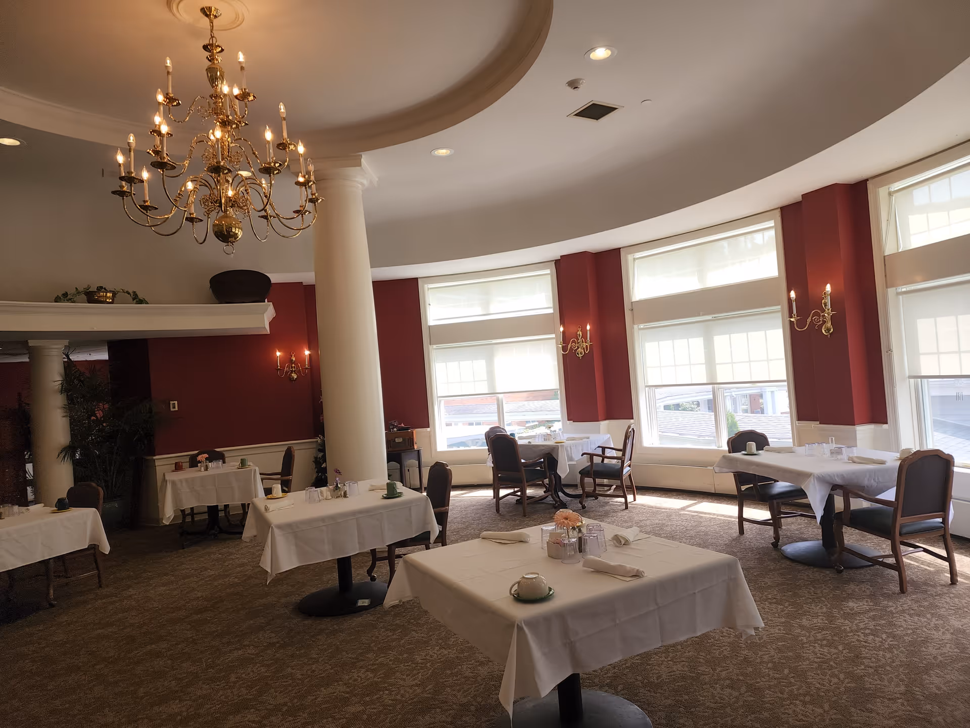 A dining room with several tables covered in white tablecloths, each set with cups, glasses, and napkins. The room features large windows with white blinds, red walls with white trim, a large white column, and an ornate chandelier hanging from the ceiling.