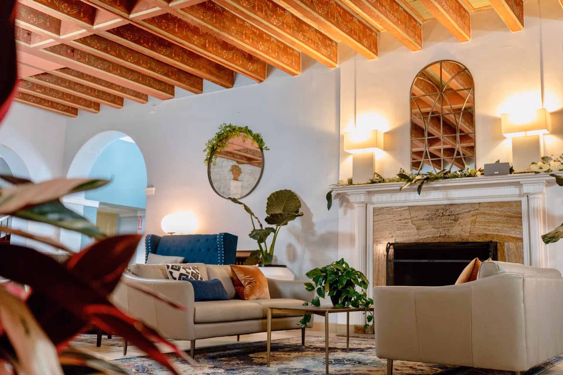 A cozy living room area with beige sofas, a blue armchair, a coffee table, and several green plants. The room features a fireplace with a decorative mirror above it, wall sconces providing warm lighting, and exposed wooden beams on the ceiling. There are arched doorways in the background and a patterned rug on the floor.