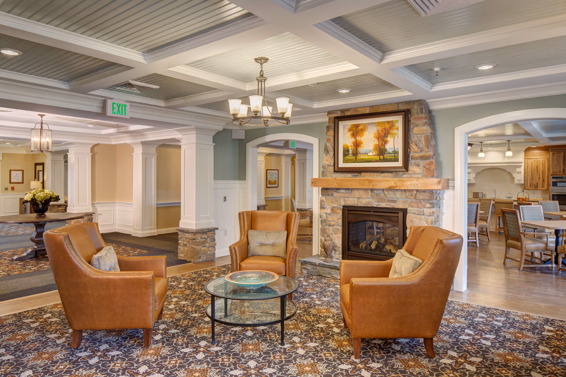 A cozy senior living facility common area with three brown leather armchairs arranged around a glass coffee table on a patterned carpet. There is a stone fireplace with a wooden mantle and a framed painting of autumn trees above it. The room features white columns, a coffered ceiling with recessed lighting, and an adjacent dining area with wooden tables and chairs.