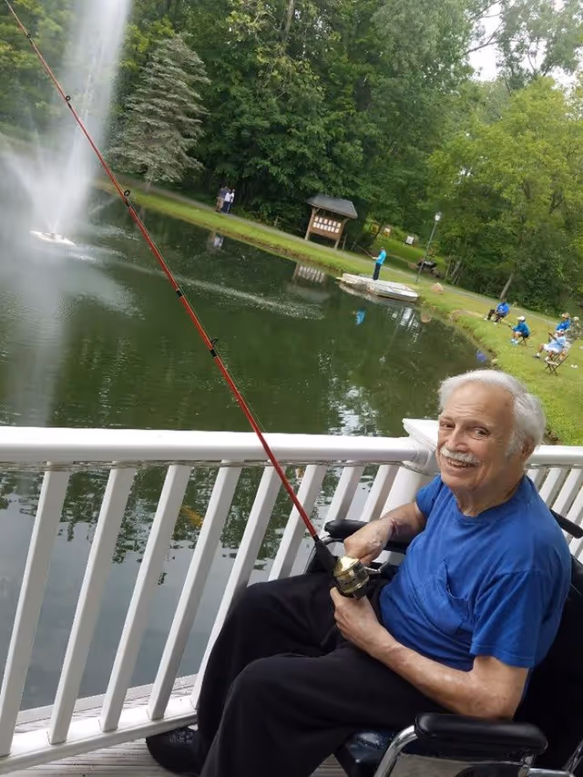 An elderly man in a wheelchair fishing with a red fishing rod on a wooden deck overlooking a pond with a water fountain. In the background, there are several people sitting and standing near the pond surrounded by green trees and grass.