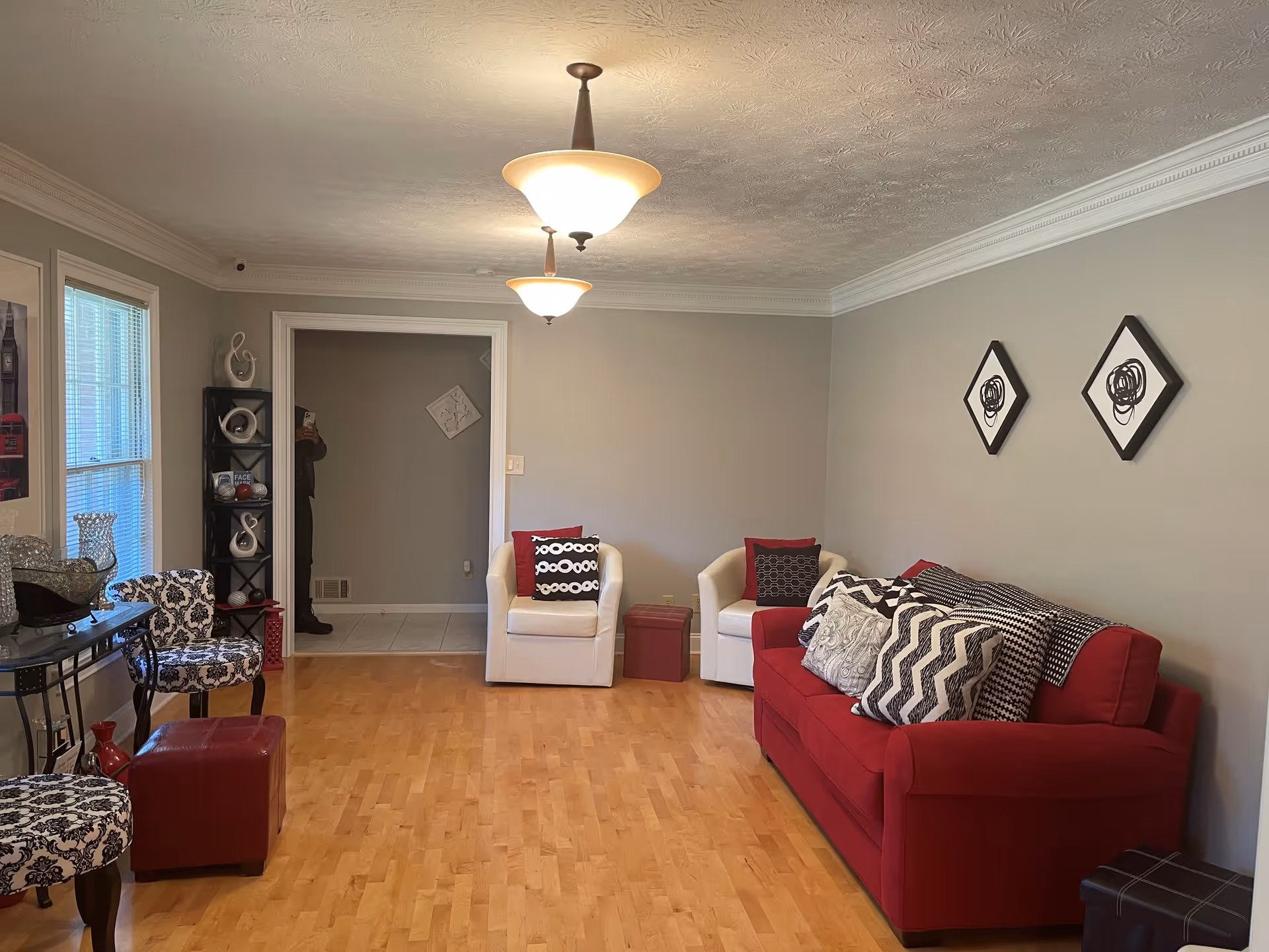A living room with light gray walls and wooden flooring. The room features a red sofa with multiple patterned cushions, two white armchairs with red and black cushions, and two patterned chairs with a small red ottoman. There are two ceiling light fixtures, a black shelving unit with decorative items, and two abstract wall art pieces in black frames. A window with blinds is on the left side, and a person taking the photo is partially visible in a doorway at the back.