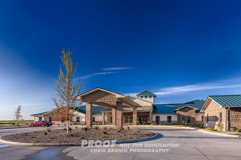 Exterior view of Carriage Crossing Senior Living of Champaign showing a single-story building with a green metal roof, a covered entrance supported by stone pillars, a circular driveway, and a few small trees and shrubs in the landscaped area under a clear blue sky.