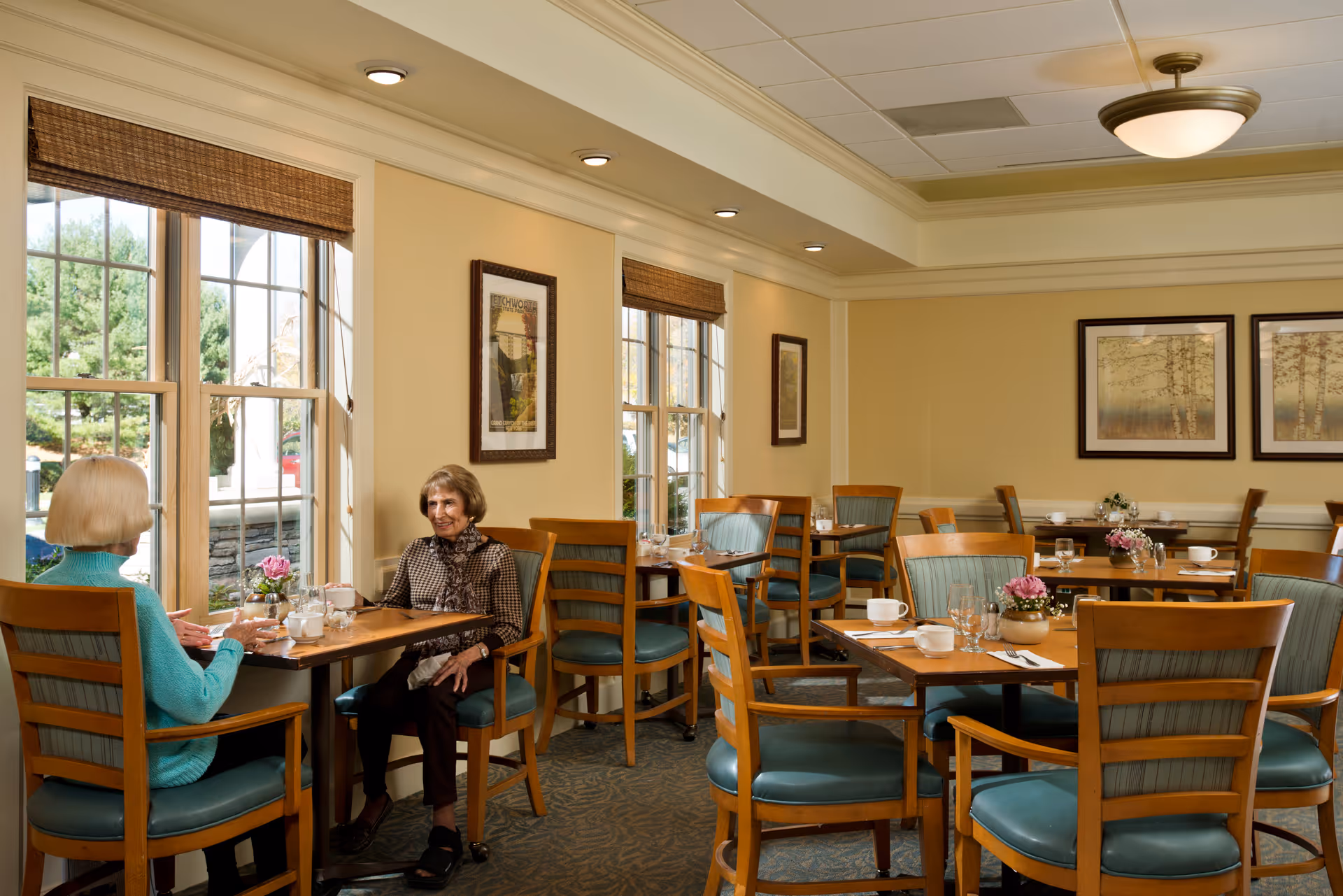 Two elderly women sitting and talking at a wooden table in a well-lit dining room with large windows, wooden chairs with teal cushions, framed artwork on the walls, and small flower arrangements on the tables.