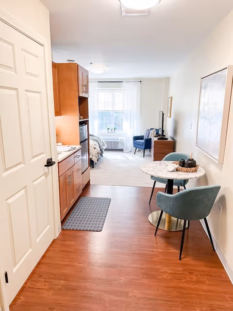 View of a bright and cozy studio apartment with wooden flooring. On the left side, there is a kitchenette with wooden cabinets, a small refrigerator, and a countertop. A small round dining table with two teal upholstered chairs is positioned on the right side. In the background, a bed is partially visible near a window with sheer white curtains, and a blue armchair is placed next to a wooden TV stand with a television on it. The walls are light-colored, and a large framed artwork hangs on the right wall.