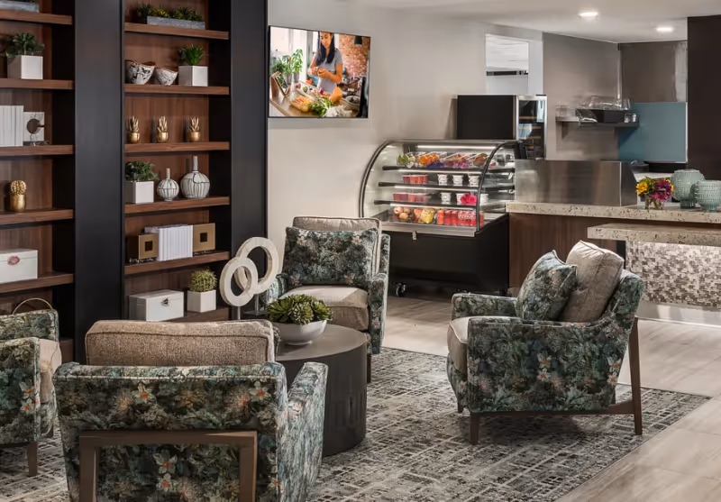 A cozy common area with four floral-patterned armchairs arranged around a round coffee table with a small plant. Behind the seating area is a wooden shelving unit with decorative items and plants. In the background, there is a refrigerated display case with food items and a counter with a mosaic tile front. A TV mounted on the wall shows a woman preparing food.