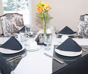 Dining table set for four with black napkins, glassware, and a vase of yellow flowers in a bright dining area.