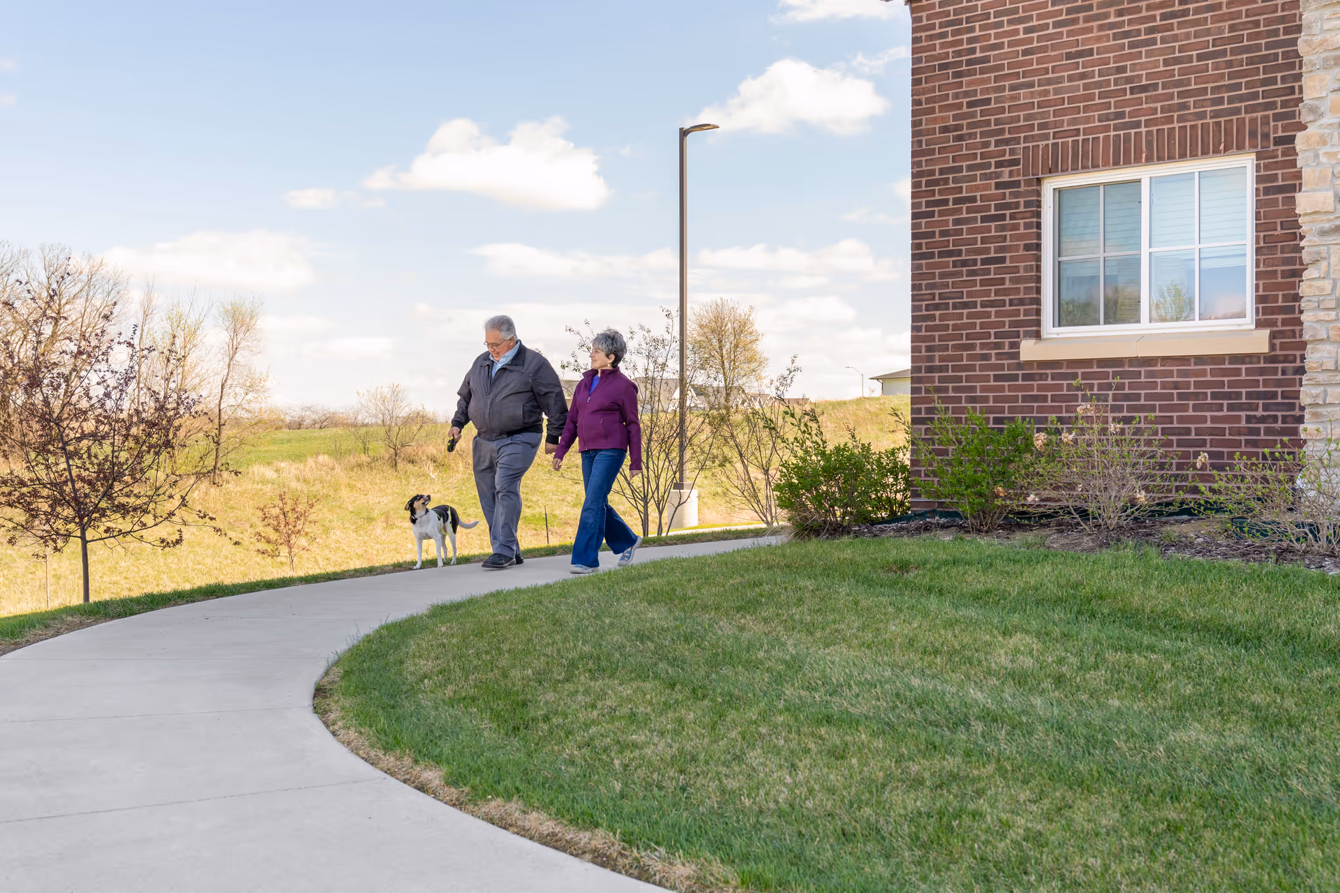 Two older adults walk a small dog along a curved sidewalk beside a brick building and grassy lawn.