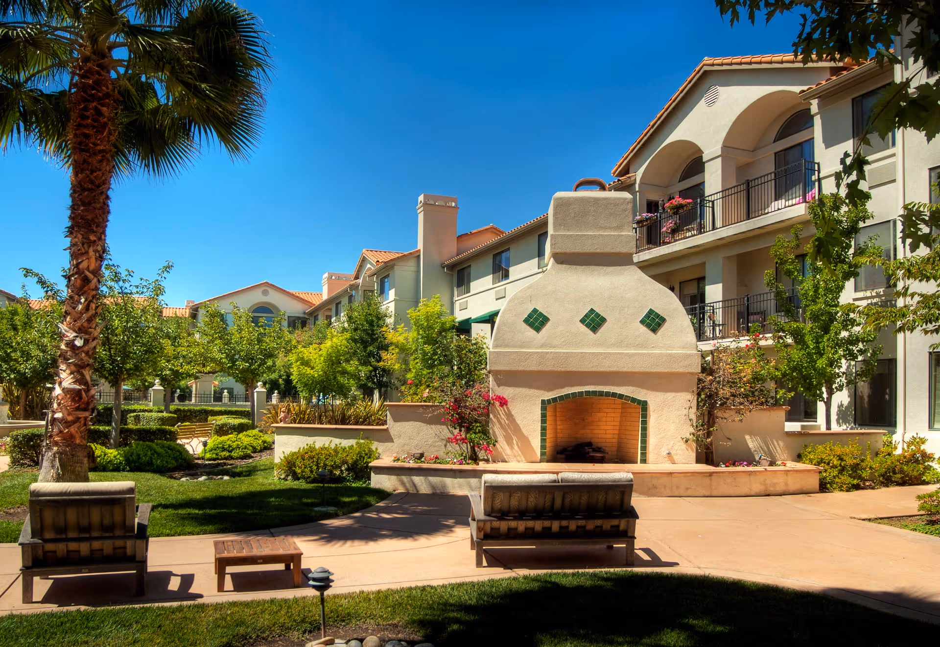 Outdoor courtyard area at Oakmont of Montecito featuring a large decorative outdoor fireplace with green tile accents, surrounded by benches and lush greenery including palm trees and other plants. The background shows a multi-story building with balconies and a clear blue sky.