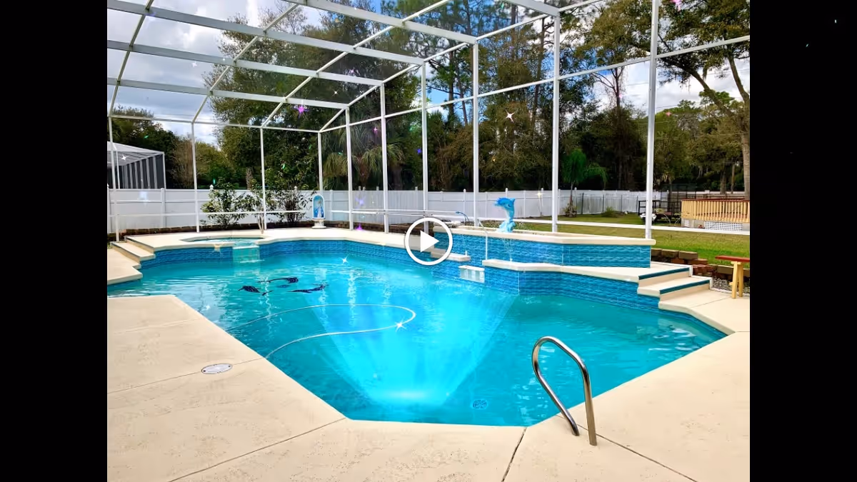 Outdoor swimming pool with clear blue water surrounded by a white fence and screened enclosure. There are steps leading into the pool, a metal handrail, and a small fountain feature in the water. Trees and greenery are visible in the background.