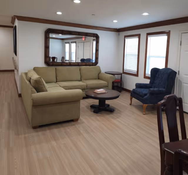 A living room with a green sectional sofa, a round wooden coffee table with books on it, a blue upholstered armchair, and a large wooden-framed mirror on the wall reflecting part of the room. The room has light wood flooring, white walls with wooden trim, two windows with blinds, and recessed ceiling lights.