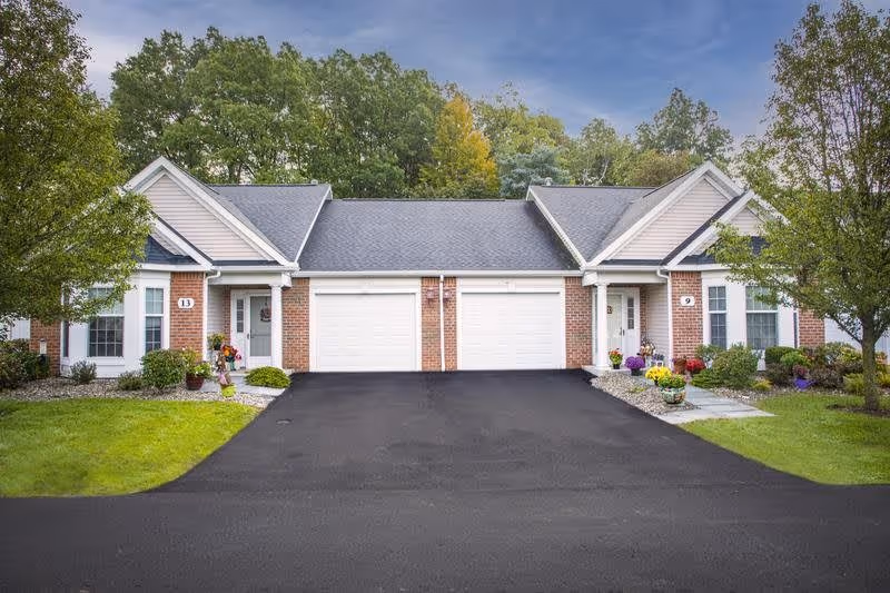 Exterior view of two attached single-story residential units with white garage doors, brick and siding walls, and small front porches. Each unit has a driveway, green lawns, trees, and flower pots near the entrances. The sky is clear with some clouds.