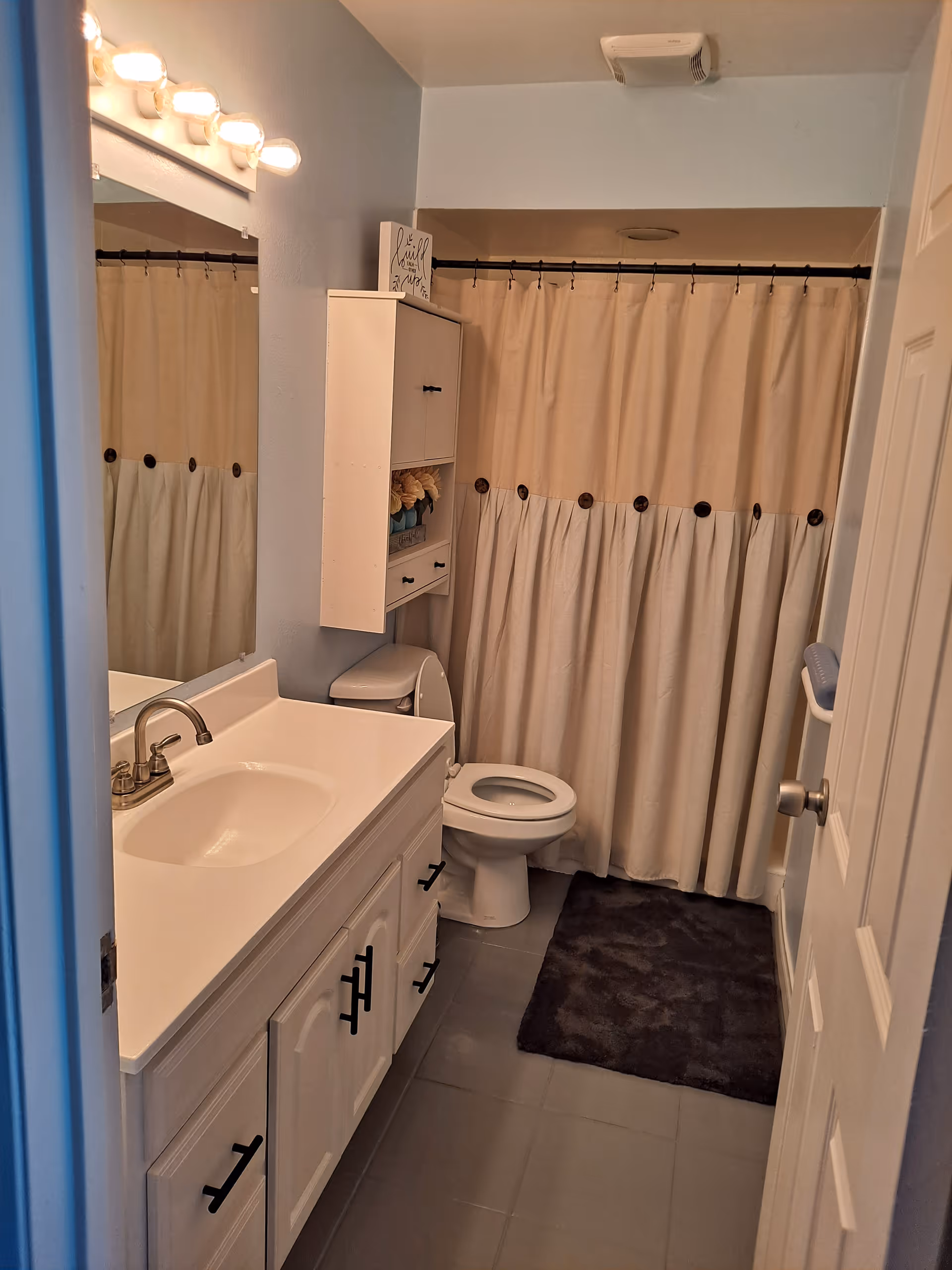 A clean bathroom with a white vanity and sink on the left, a toilet next to it, and a shower with a beige and white curtain in the background. There is a dark gray bath mat on the floor and a white cabinet mounted on the wall above the toilet. The bathroom has light blue walls and a mirror above the sink with four light bulbs.