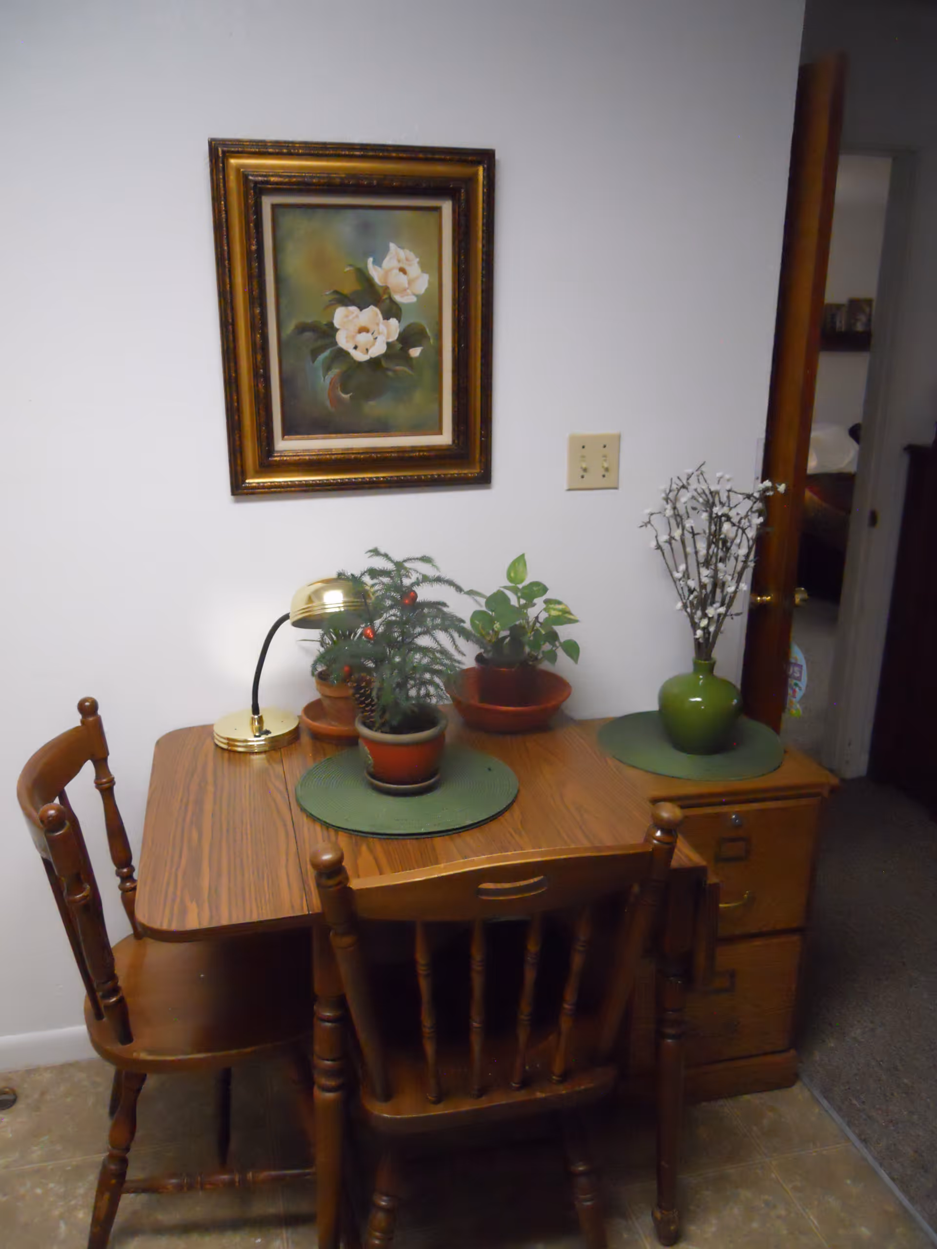 A small wooden table with two wooden chairs in a corner of a room. On the table are three potted plants, a small gold desk lamp, and green placemats. A framed painting of white flowers hangs on the wall above the table. To the right, there is a wooden filing cabinet with a green vase holding white flowers. An open doorway leads to another room.