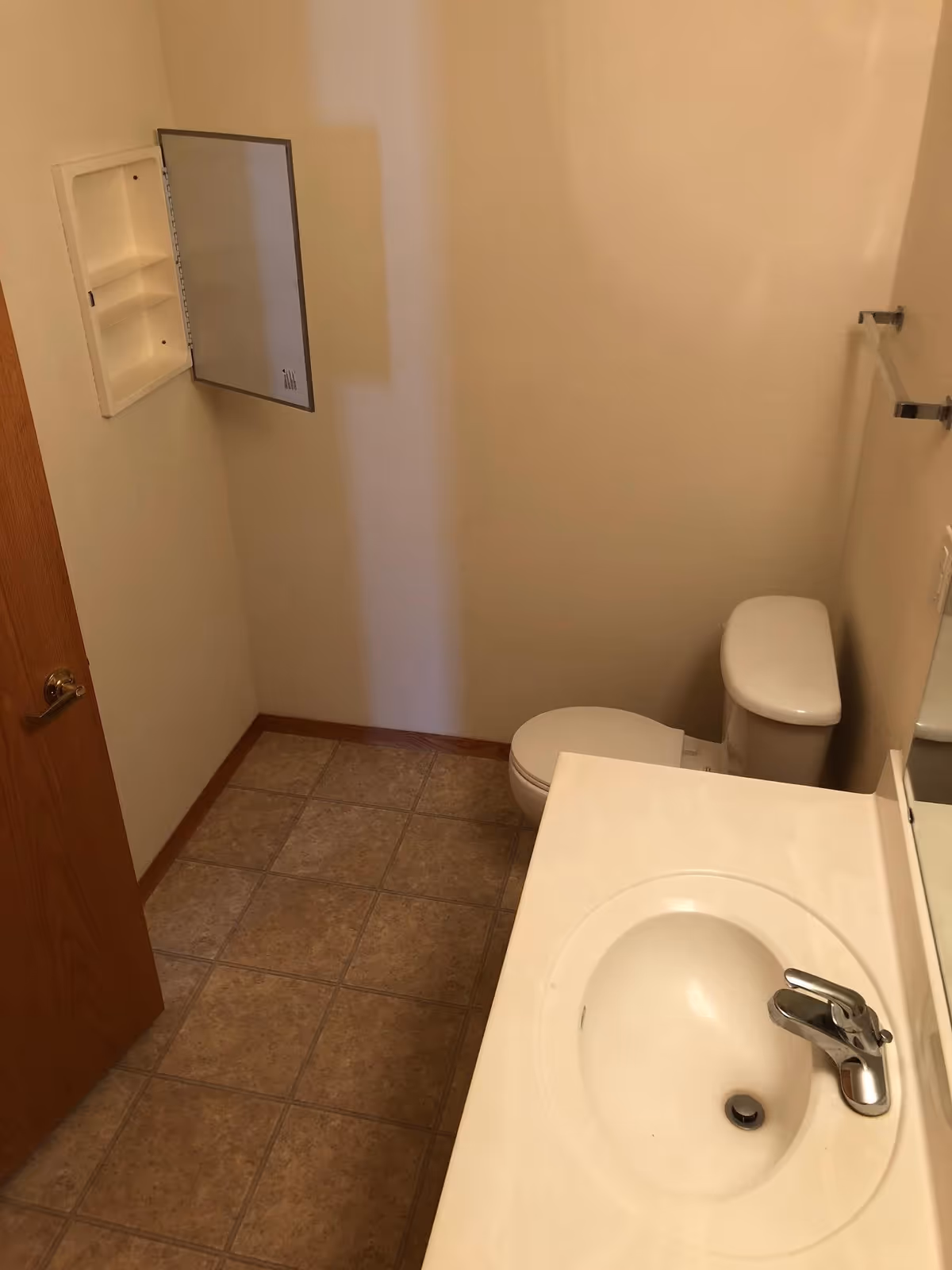 Small bathroom with a sink and countertop in the foreground, a toilet in the back right, an open recessed medicine cabinet on the left wall, and tiled floor.