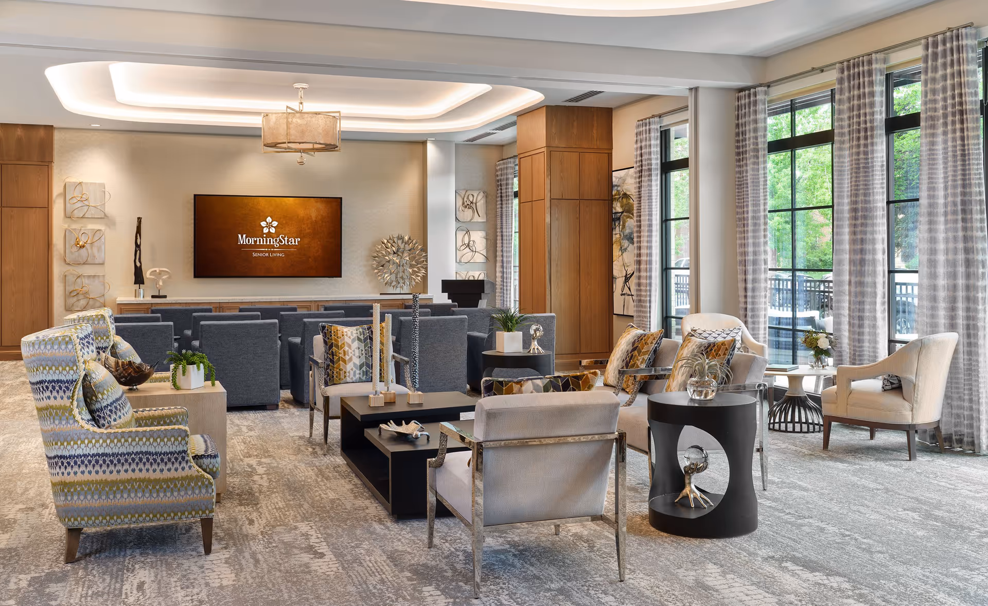 A spacious and well-lit living room area in MorningStar Assisted Living & Memory Care at Observatory Park featuring modern and comfortable seating including patterned armchairs, beige chairs, and gray sofas. The room has large windows with patterned curtains allowing natural light to fill the space. Decorative elements such as plants, sculptures, and abstract wall art add to the inviting ambiance. A large screen on the far wall displays the MorningStar Senior Living logo.