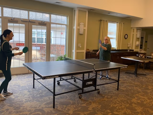 Two women playing table tennis in a senior living community common room with large windows and a table in the background.