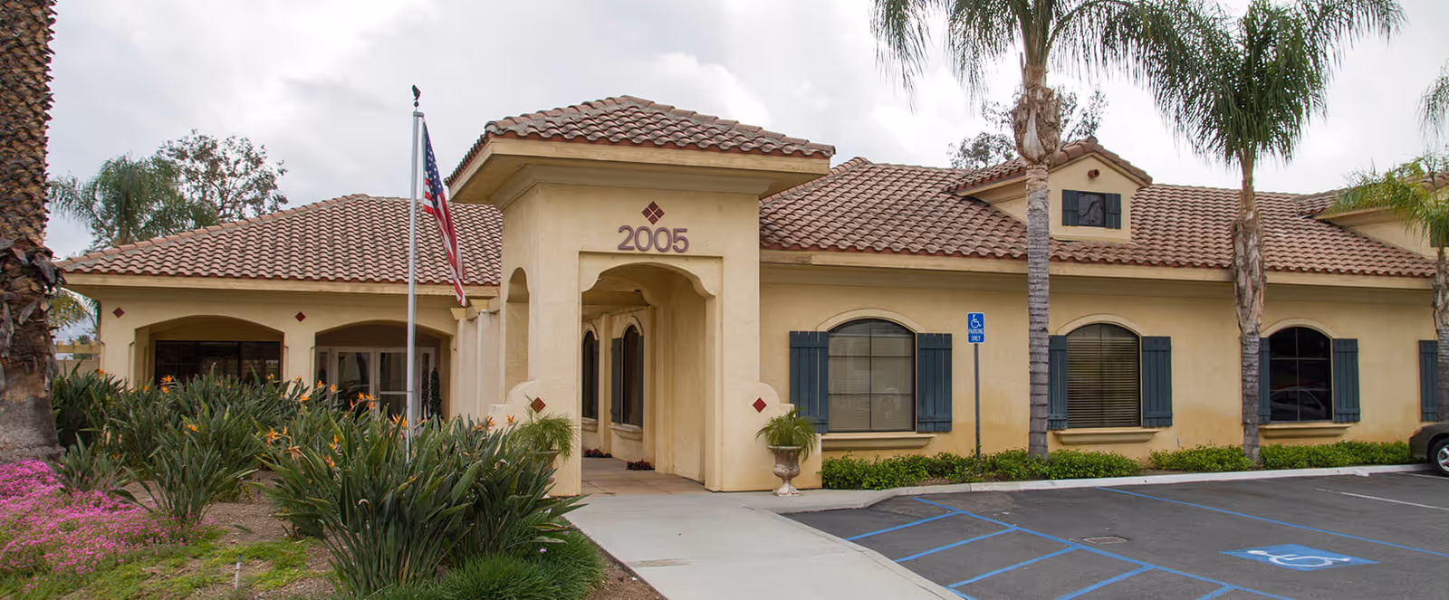 Exterior view of a single-story building with beige stucco walls and a red tile roof, featuring the number 2005 above the entrance. There are palm trees, green shrubs, and pink flowers in the landscaped area near the sidewalk. An American flag is flying on a flagpole near the entrance. A parking lot with marked handicapped parking spaces is visible in front of the building.