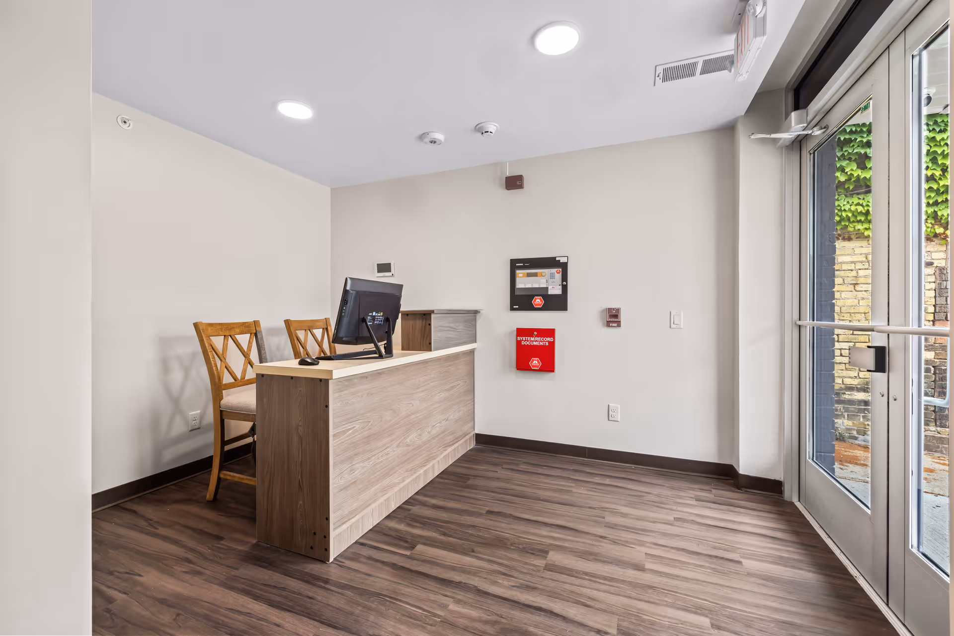 Reception area with a wooden desk, two wooden chairs with cushioned seats, a computer monitor on the desk, and a glass door leading outside. The walls are white, and there is a fire alarm system and a red emergency box mounted on the wall.