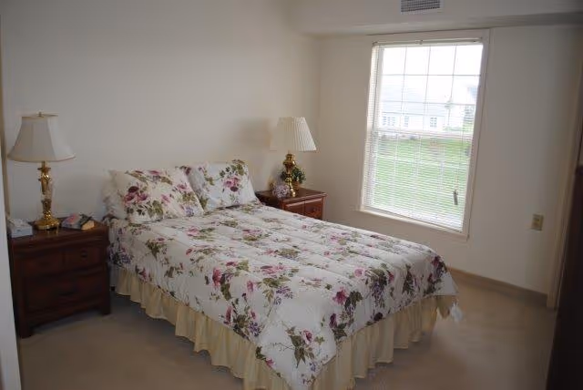 A neatly made bed with floral bedding flanked by two wooden nightstands and lamps in a bright bedroom with a window.
