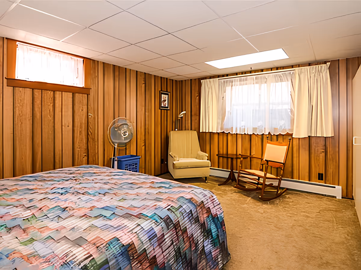 A bedroom with wood-paneled walls and a drop ceiling. The room features a bed with a colorful quilt, a beige armchair, a wooden rocking chair, a small wooden side table, and a standing fan on a blue crate. There are two windows with sheer white curtains allowing natural light into the room.