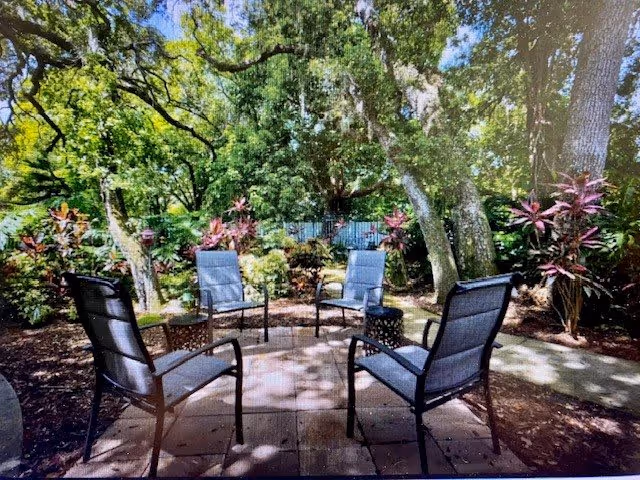 A shaded outdoor patio with five metal-framed chairs arranged in a circle on paving stones surrounded by lush trees and plants.