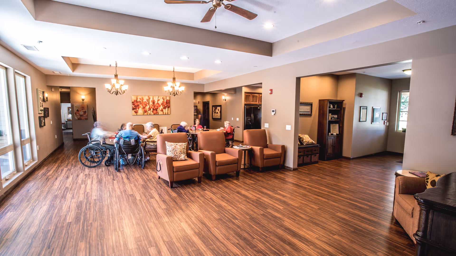 A spacious senior living common area with wooden flooring, several armchairs, and a group of elderly people sitting around a table near large windows. The room is well-lit with ceiling lights and chandeliers, and decorated with wall art and furniture.